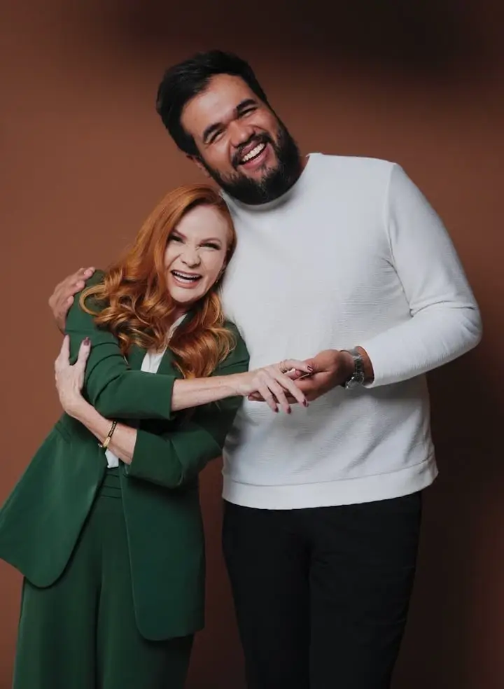 Cátia Damasceno, de terno verde, sorrindo ao lado de homem de suéter branco, em retrato por Victor Moura