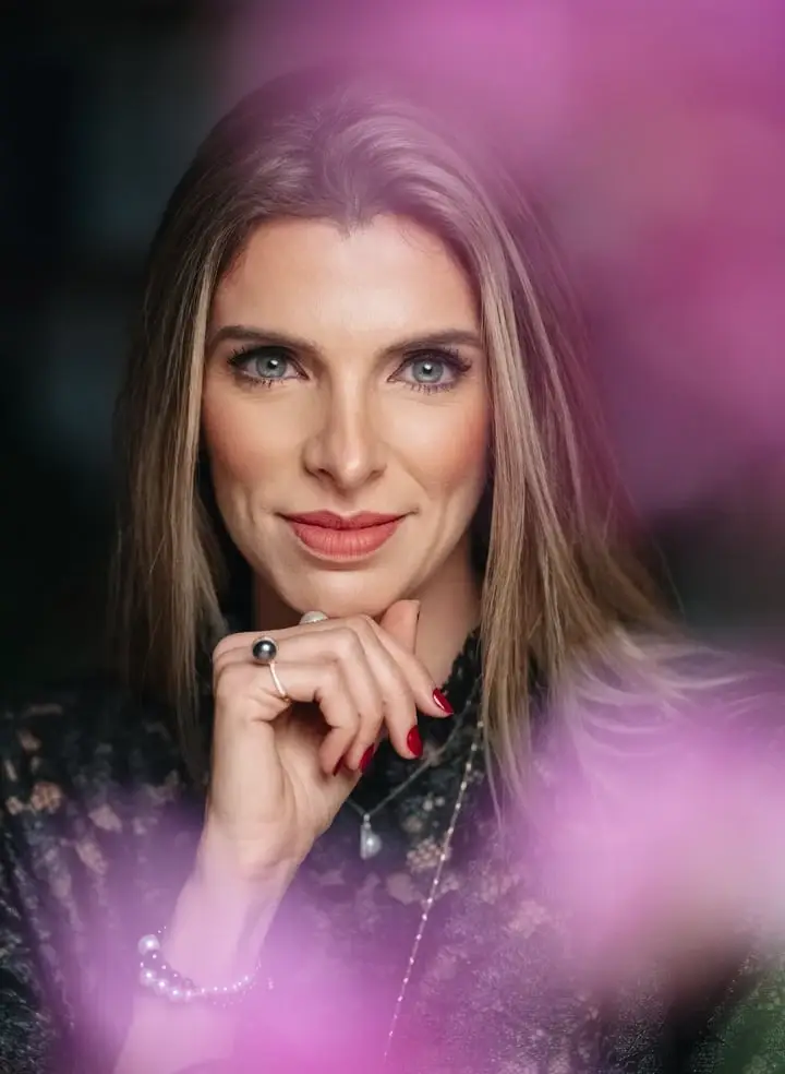 Mariana Parada com a mão no queixo, entre flores rosadas, em retrato profissional por Victor Moura