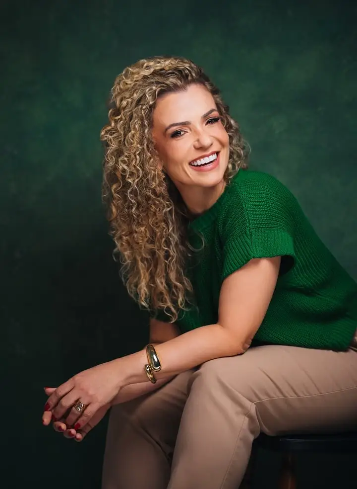 Mulher de cabelo cacheado e blusa verde, sentada e sorrindo, em retrato por Victor Moura